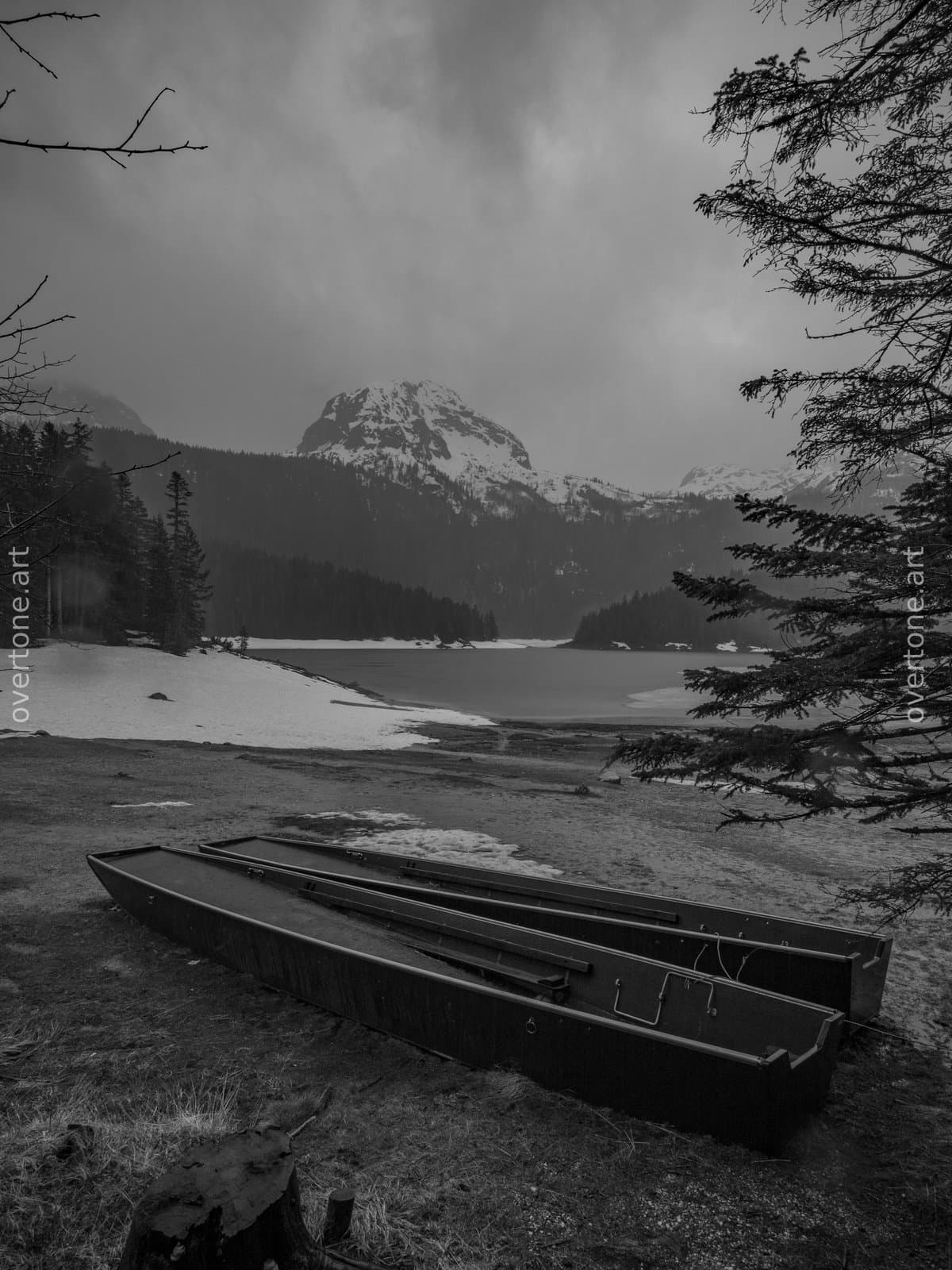 Two wooden rowboats rest on the shore of Black Lake (Crno Jezero) in Durmitor National Park, Montenegro, beside a partially frozen lake with snow-covered mountains in the background. - Wild Places