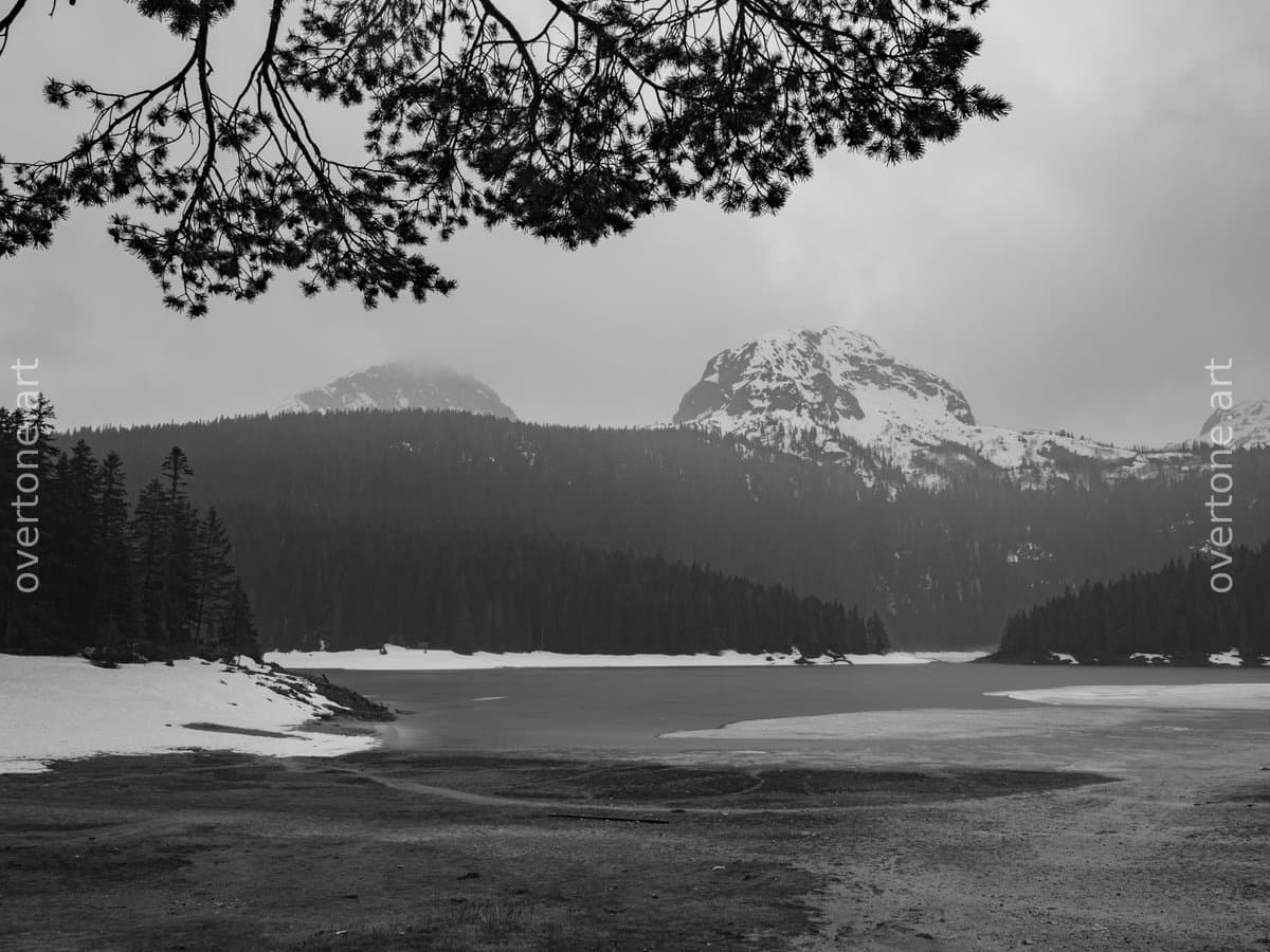 Black-and-white photograph of Black Lake (Crno Jezero) in Durmitor National Park, Montenegro, with a quiet shoreline, melting snow, evergreen forest, and snow-covered mountain peaks. - Wild Places